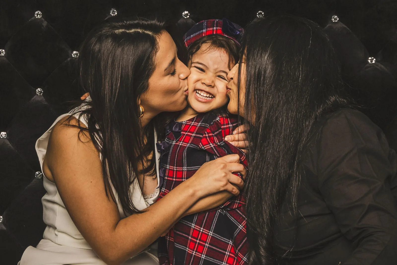 Both parents kissing their daughter's cheeks against a dark tufted backdrop
