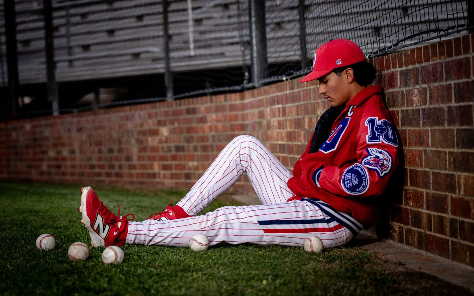 Senior in uniform and letter jacket seated against a brick dugout wall