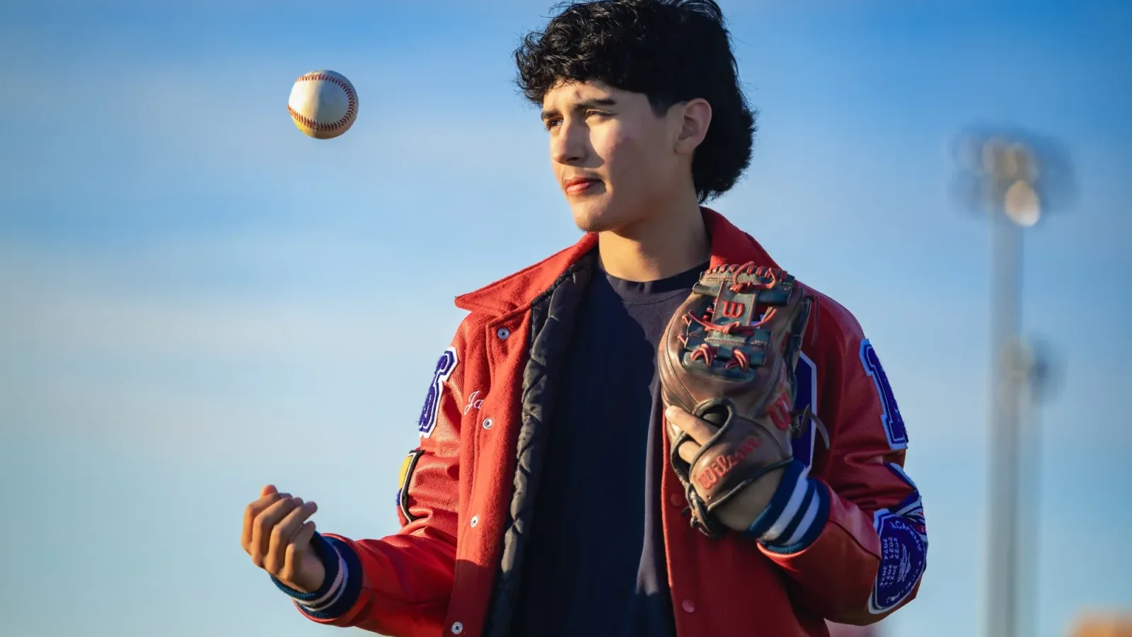 High school senior in a red letterman jacket tossing a baseball, glove on his hand, against an open sky