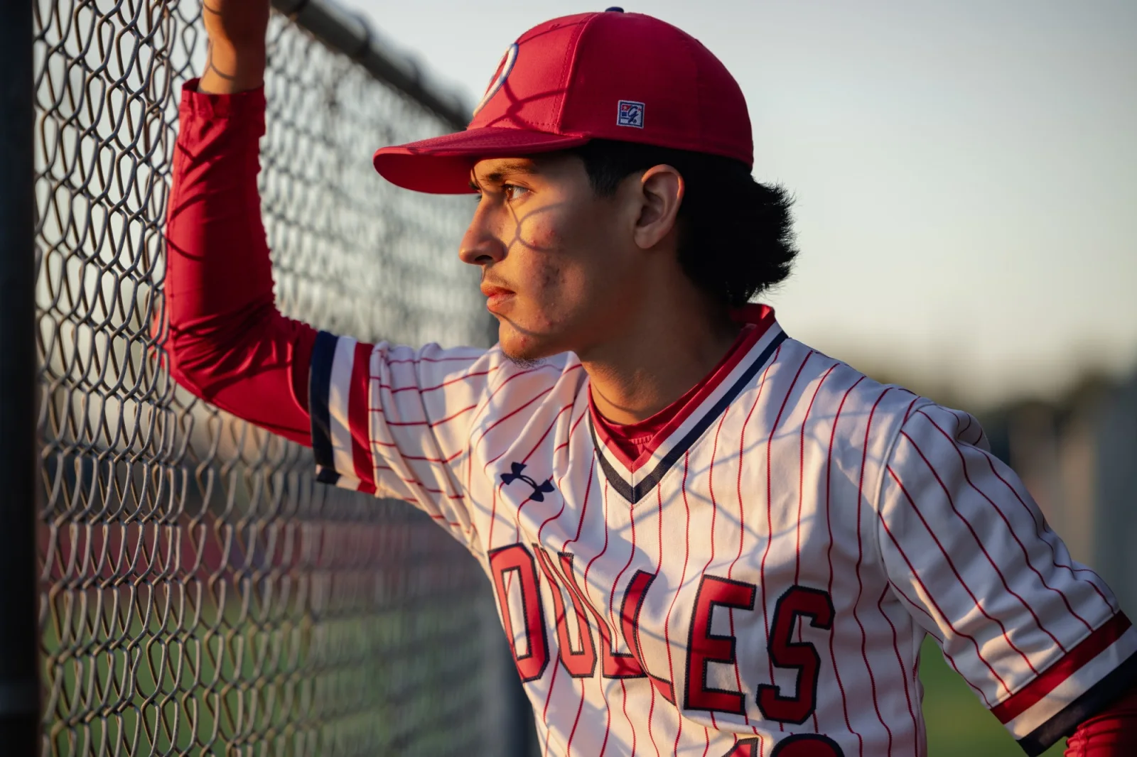 Senior in his baseball uniform leaning against the outfield fence at golden hour
